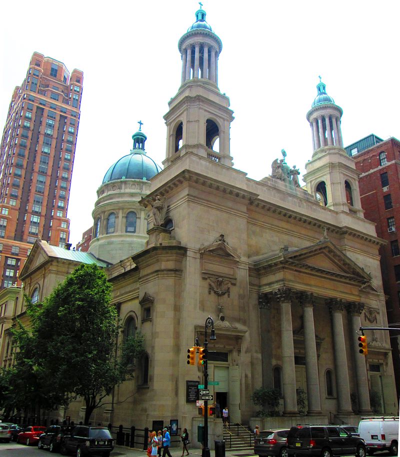 A stone building with a pediment and columns on the main entrance, two towers with green rounded tops and columns, and a dome at the rear