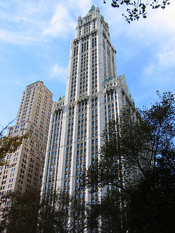 Color photo of the Woolworth Building with trees in the foreground and a tall but significantly shorter building to the left