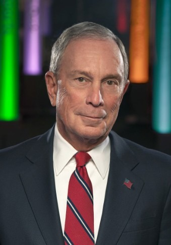 profile of Michael Bloomberg, in a grey suit, and red necktie with blue stripe, with wearing an American flag pin