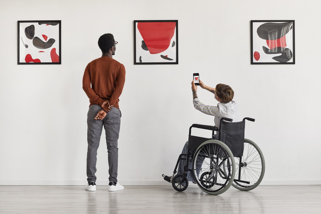 Back view portrait of young woman using wheelchair taking photo of artwork while visiting accessible museum