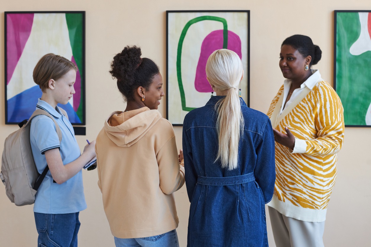 Diverse group of teenage schoolchildren listening to female tour guide in modern art gallery